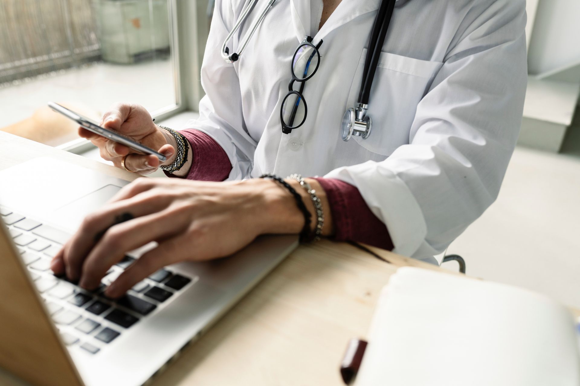 Doctor in white coat using laptop and cell phone, stethoscope around neck, glasses hanging.