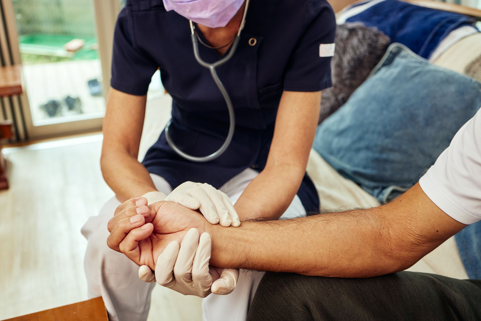 A healthcare worker checks a patient's wrist pulse with a stethoscope in a home setting.