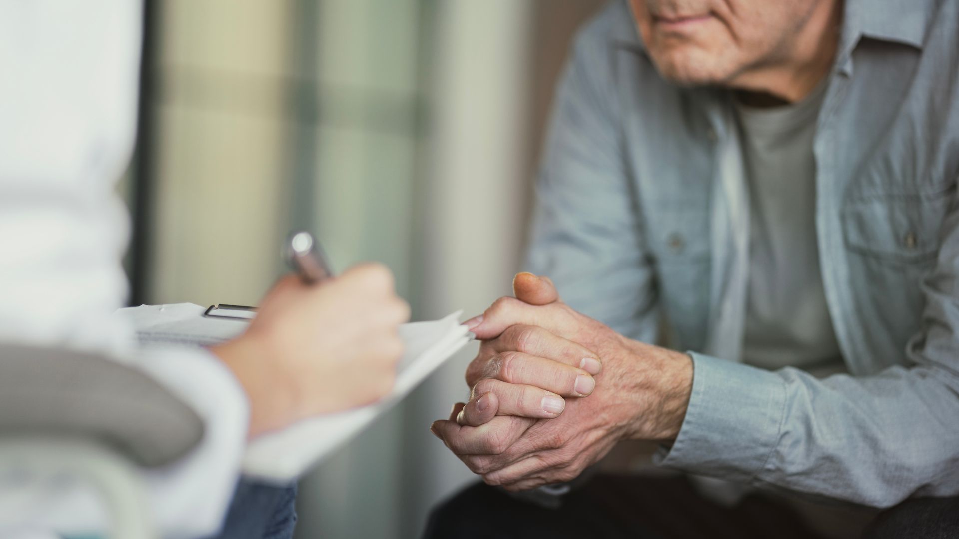 Person sitting with hands clasped, listening to someone writing on a clipboard. Person sitting with hands clasped, listening to someone writing on a clipboard.