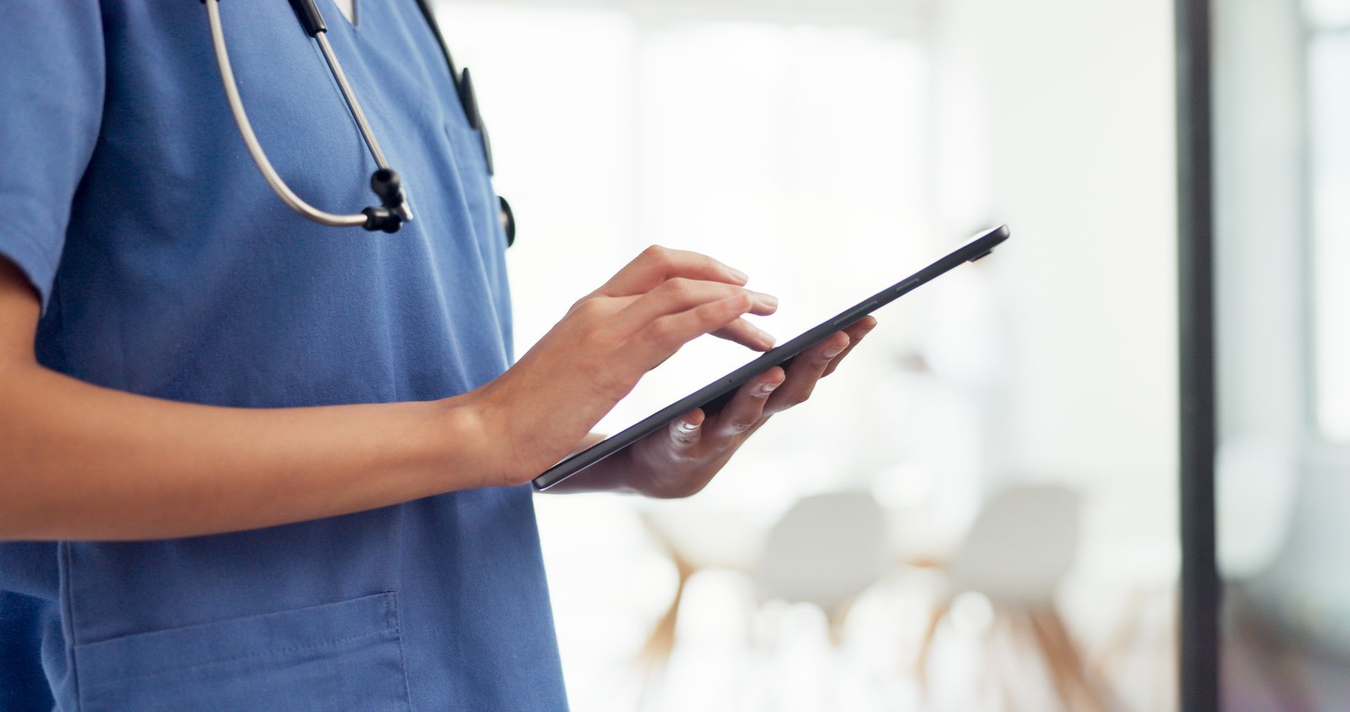 Healthcare worker in blue scrubs using a tablet, stethoscope around neck.