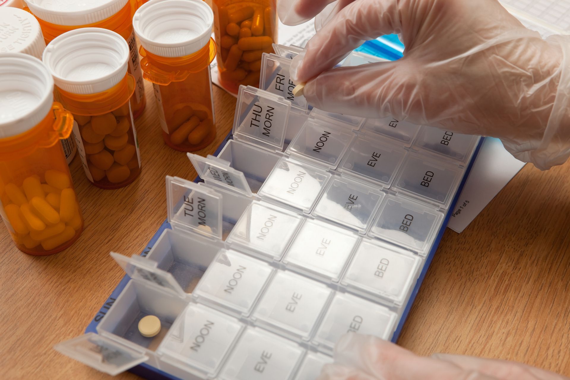 Person filling a pill organizer with medication from orange prescription bottles, wearing gloves.