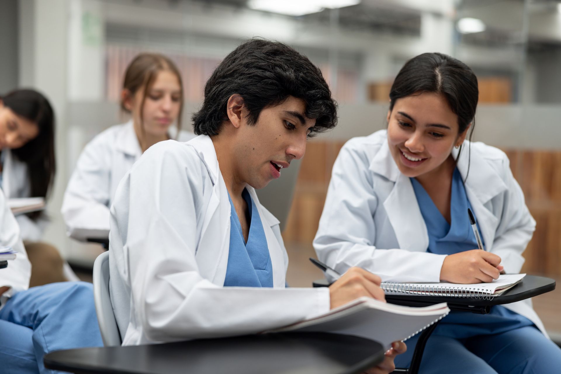 Two medical students in white coats study together in a classroom. Two medical students in white coats study together in a classroom.