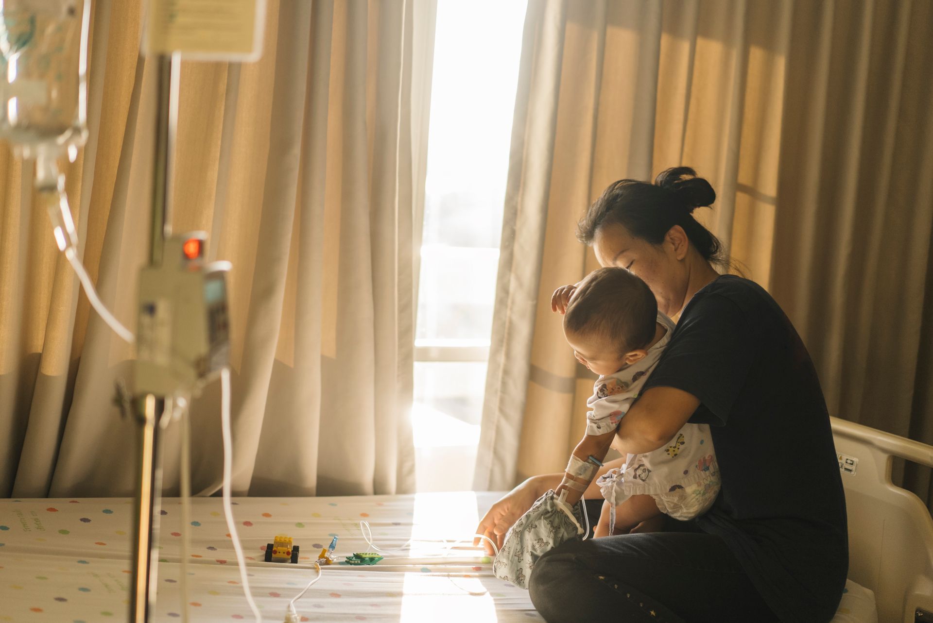 Woman holding a child in a hospital room, sunlight streams through a window. An IV drip bag hangs nearby.