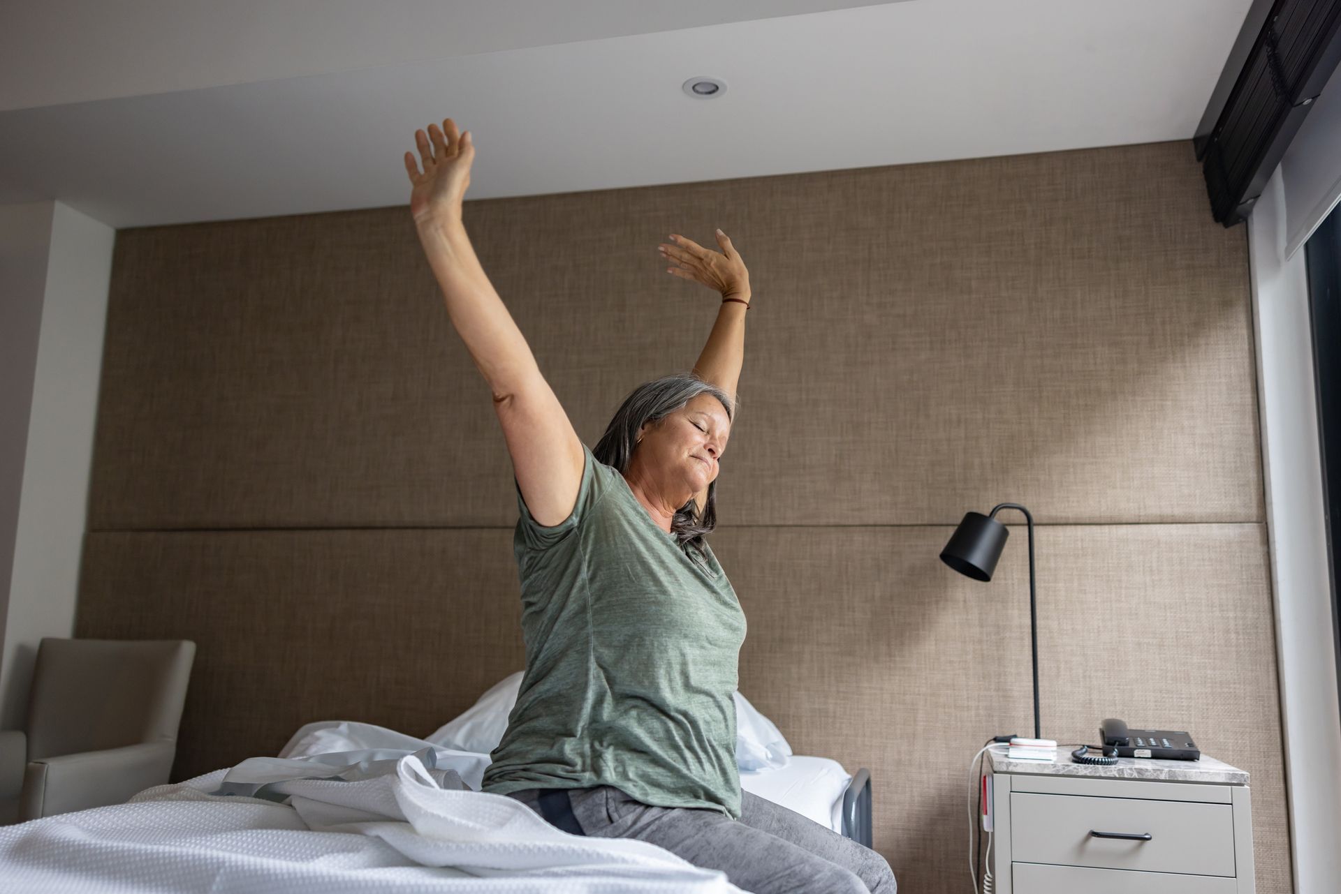 Woman sitting up in bed, stretching arms upward. Beige headboard in background. Woman sitting up in bed, stretching arms upward. Beige headboard in background.