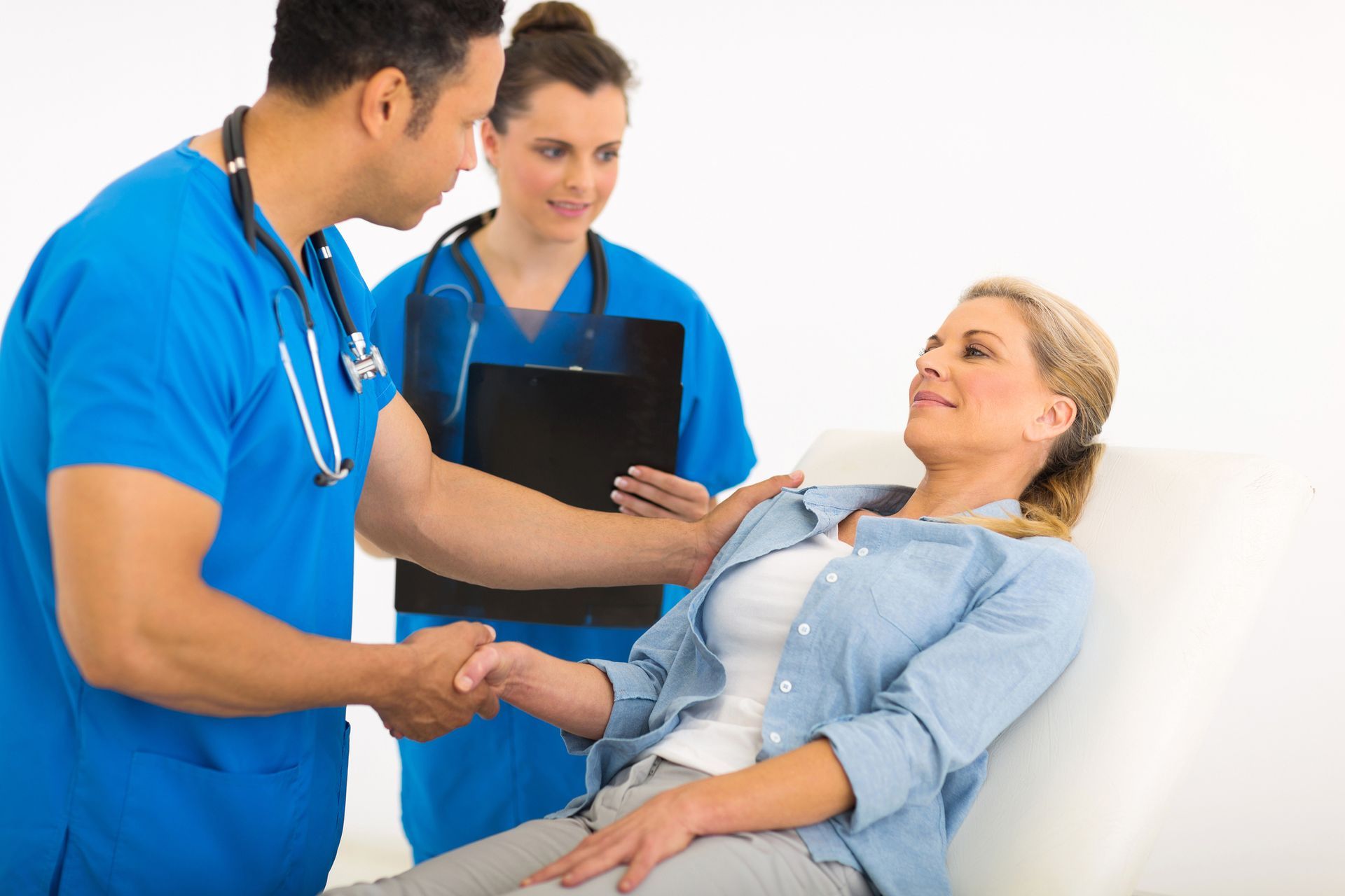 Medical professionals assisting a patient lying on an examination table. One doctor shakes the patient’s hand; a nurse holds a clipboard.