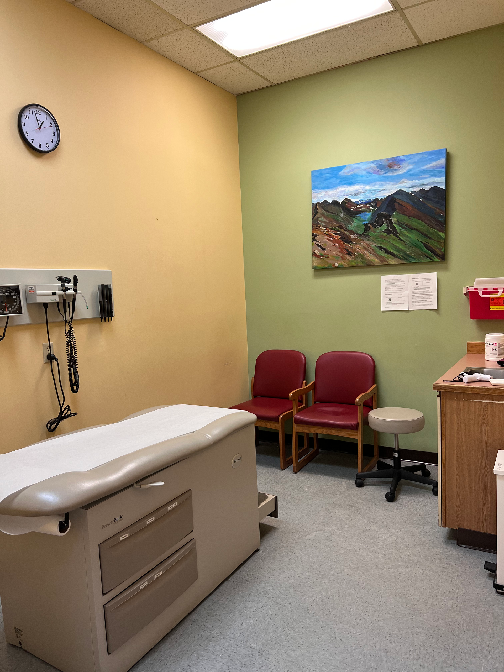 Medical exam room with examination table, chairs, and artwork on the wall.