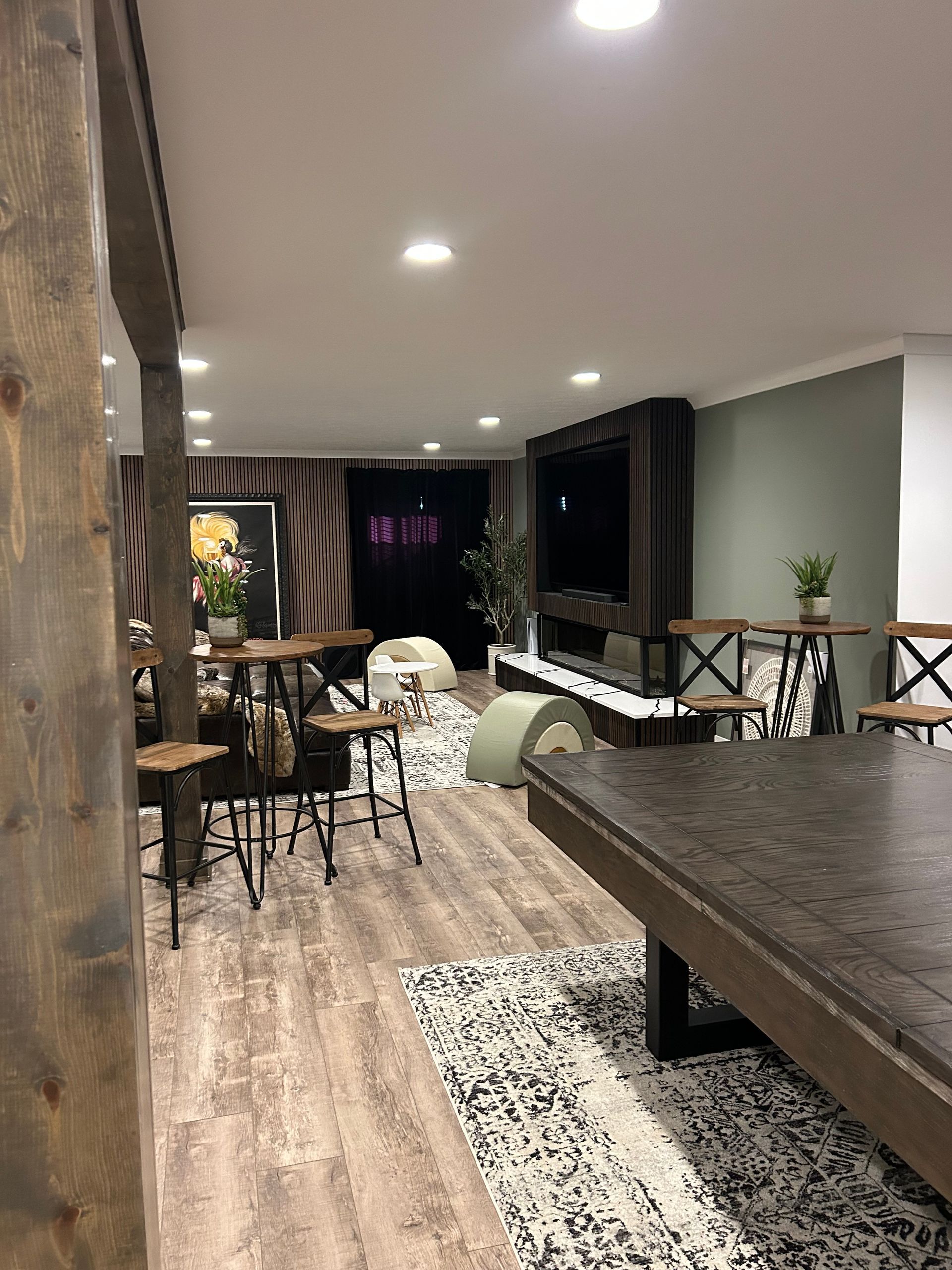 Modern open-plan basement with wood floors, recessed lights, bar stools, and a large dark dining table