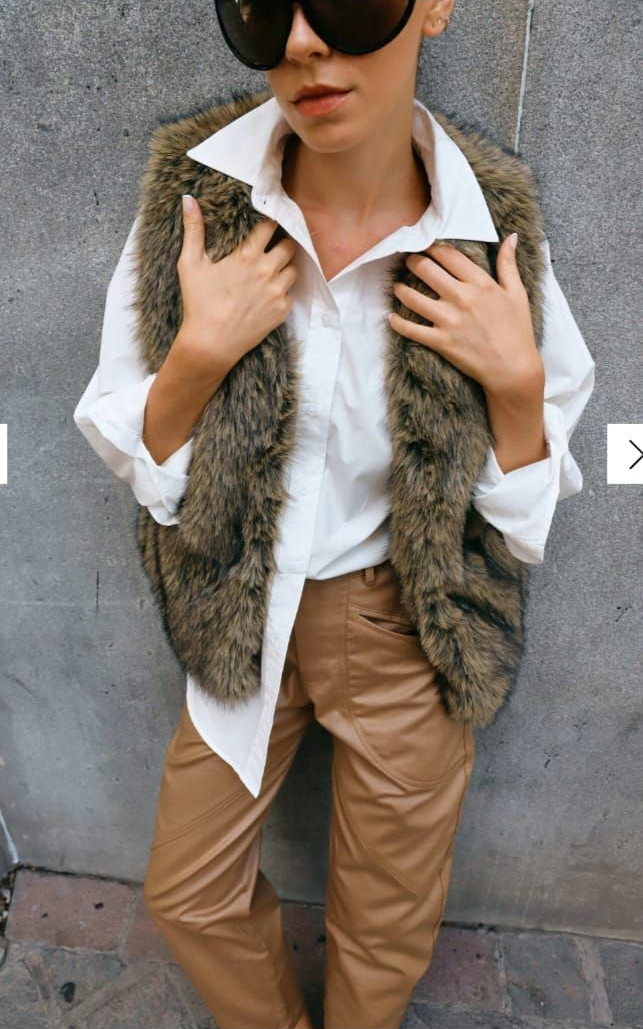 Persona de pie junto a un muro de piedra, vestida con camisa blanca, pantalones marrones y chaleco de piel.