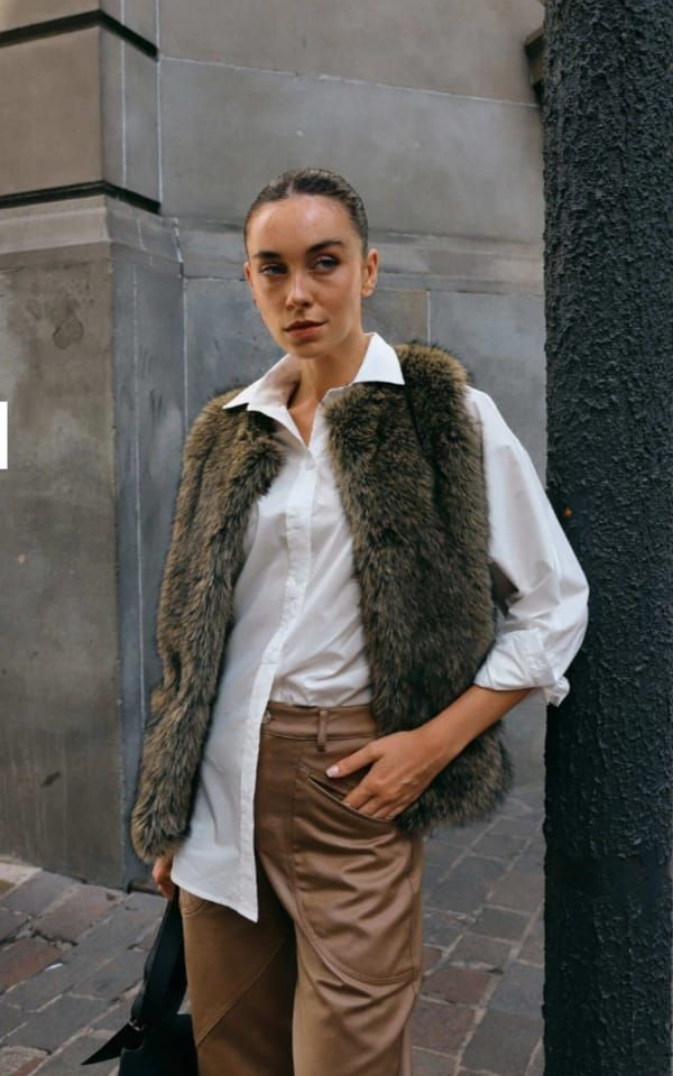 Persona de pie al aire libre, vestida con camisa blanca y pantalón marrón, y con un chaleco de piel.