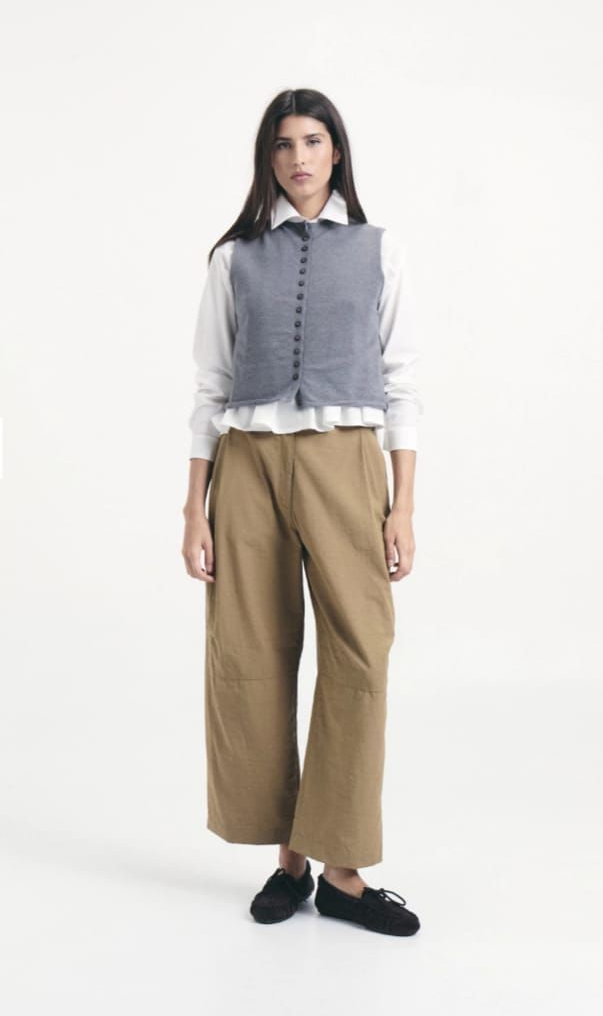 Mujer con blusa gris y pantalones anchos color canela, de pie sobre un fondo blanco.