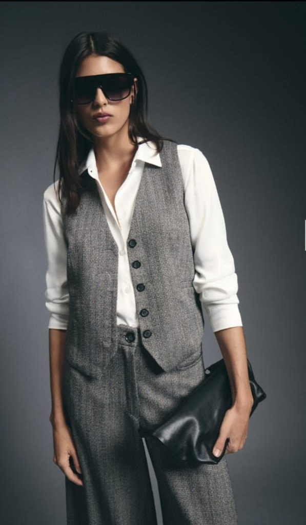 Modelo con camisa blanca, chaleco gris y pantalones anchos, posando frente a un fondo gris de estudio.