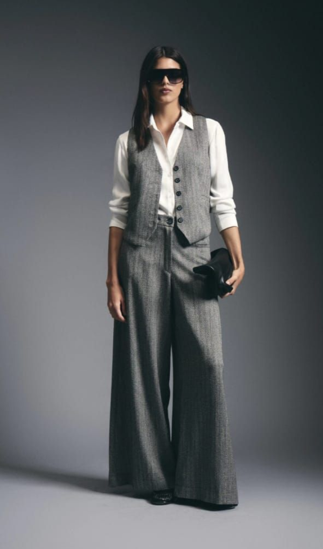 Modelo con camisa blanca, chaleco gris y pantalones anchos, posando frente a un fondo gris de estudio.