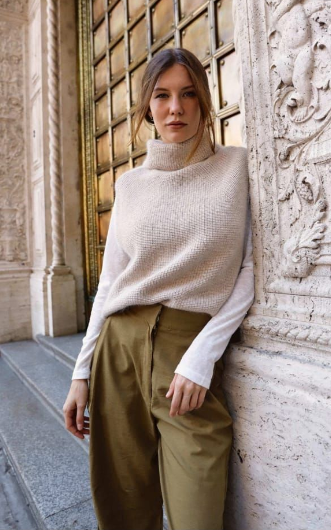 Persona con jersey de cuello alto color crema y pantalones verde oliva de pie junto a una pared de piedra tallada.