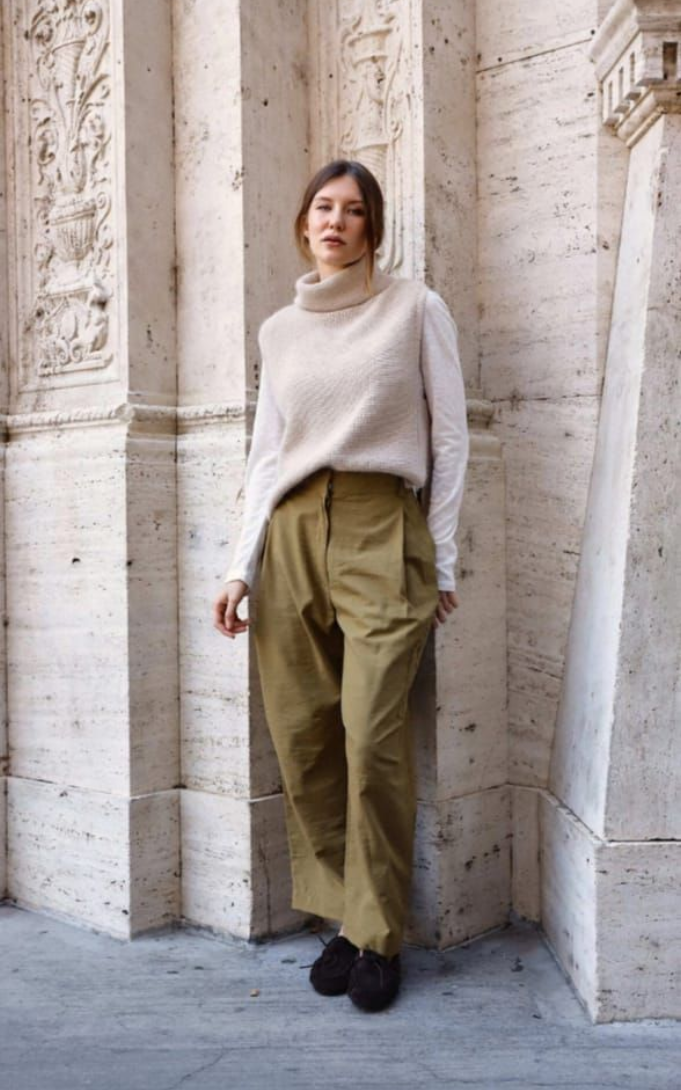 Persona con jersey de cuello alto color crema y pantalones verde oliva de pie junto a una pared de piedra tallada.