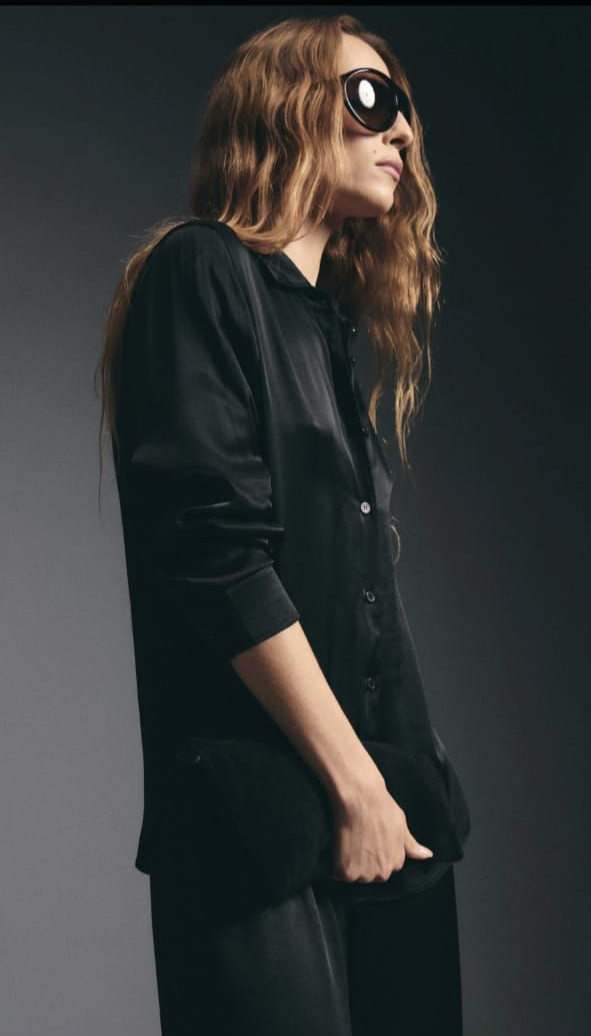 Mujer vestida de negro y con gafas de sol caminando frente a un fondo de estudio gris.