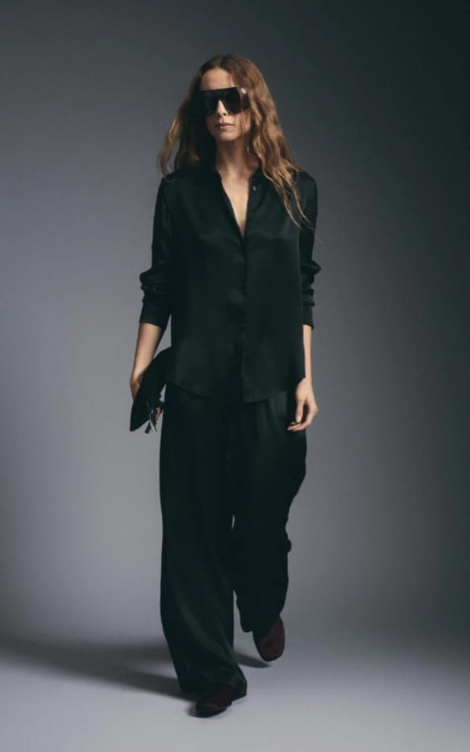 Mujer vestida de negro y con gafas de sol caminando frente a un fondo de estudio gris.