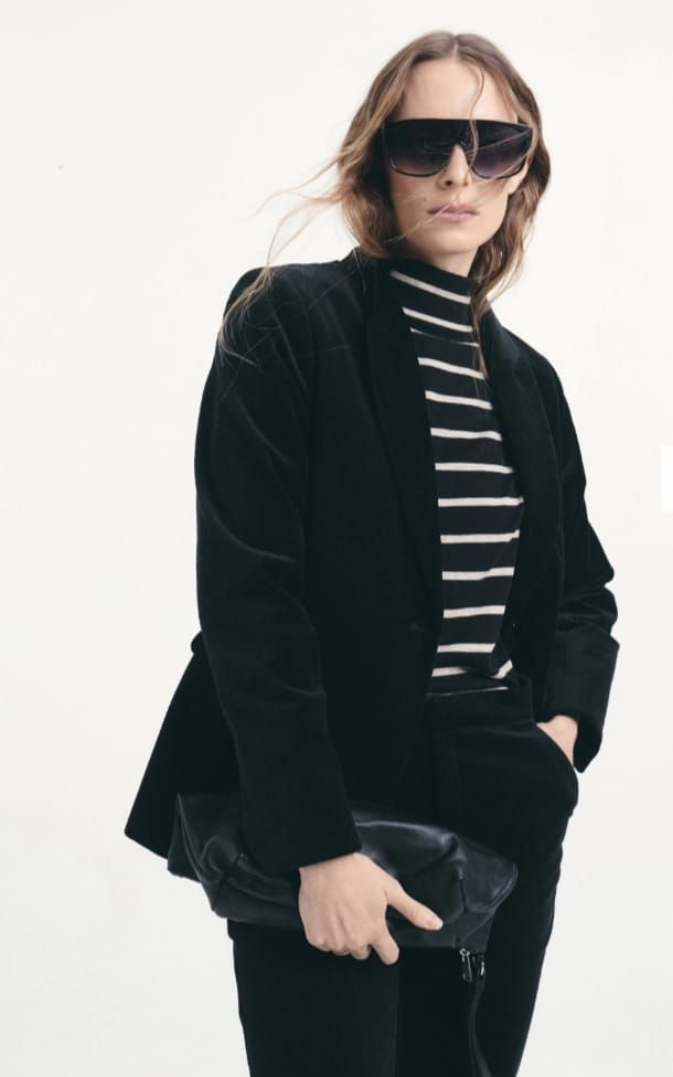 Persona con chaqueta negra y jersey de cuello alto a rayas, con gafas de sol, sentada sobre un fondo blanco.