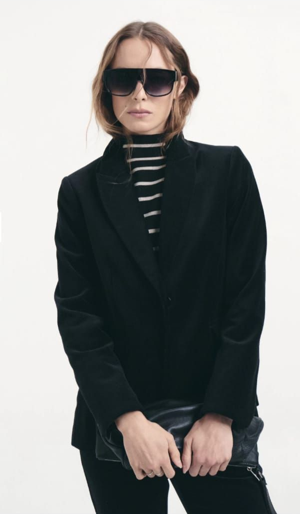 Persona con chaqueta negra y jersey de cuello alto a rayas, con gafas de sol, sentada sobre un fondo blanco.