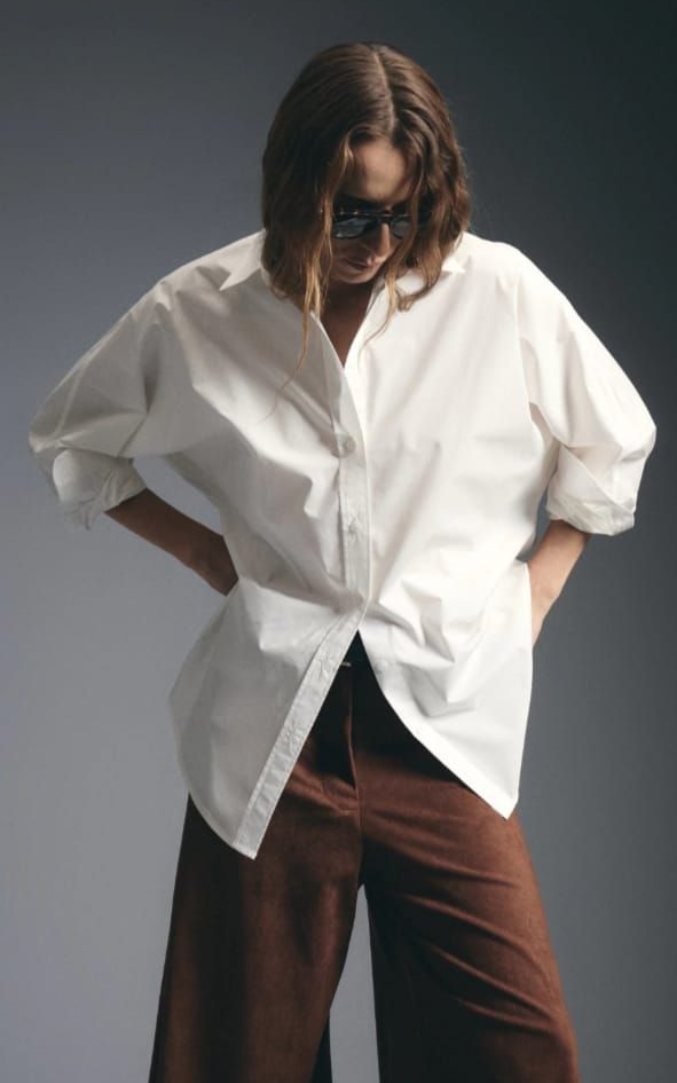 Persona con camisa blanca y pantalones marrones mirando hacia abajo con las manos en los bolsillos sobre un fondo gris.