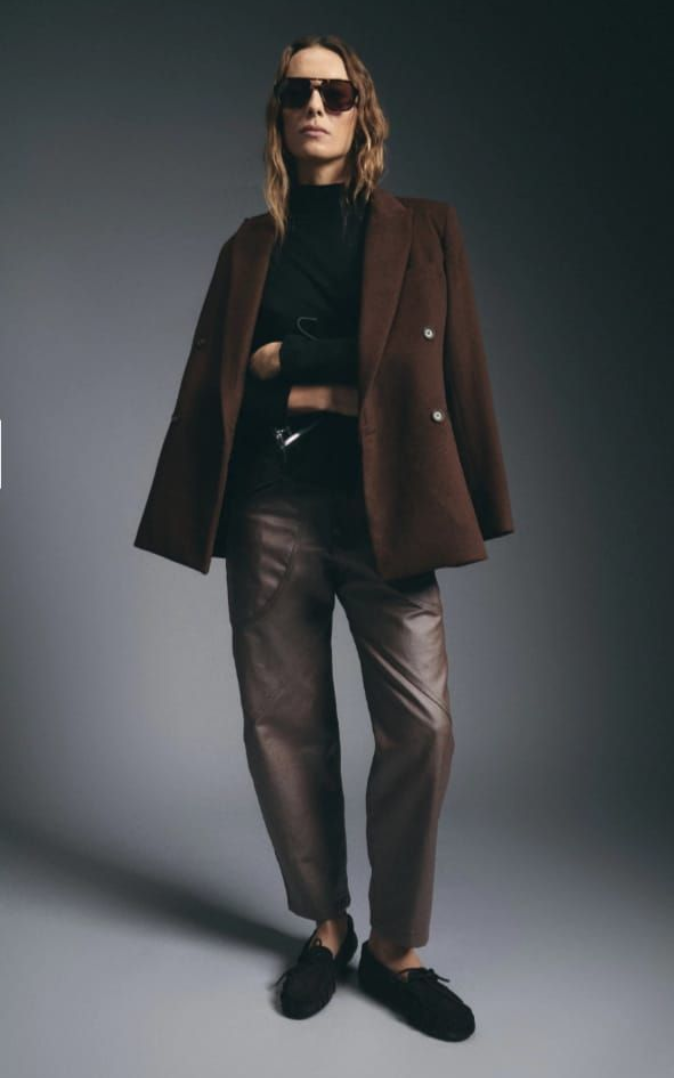 Persona con chaqueta oscura y pantalones de satén caminando de perfil contra un fondo gris de estudio.