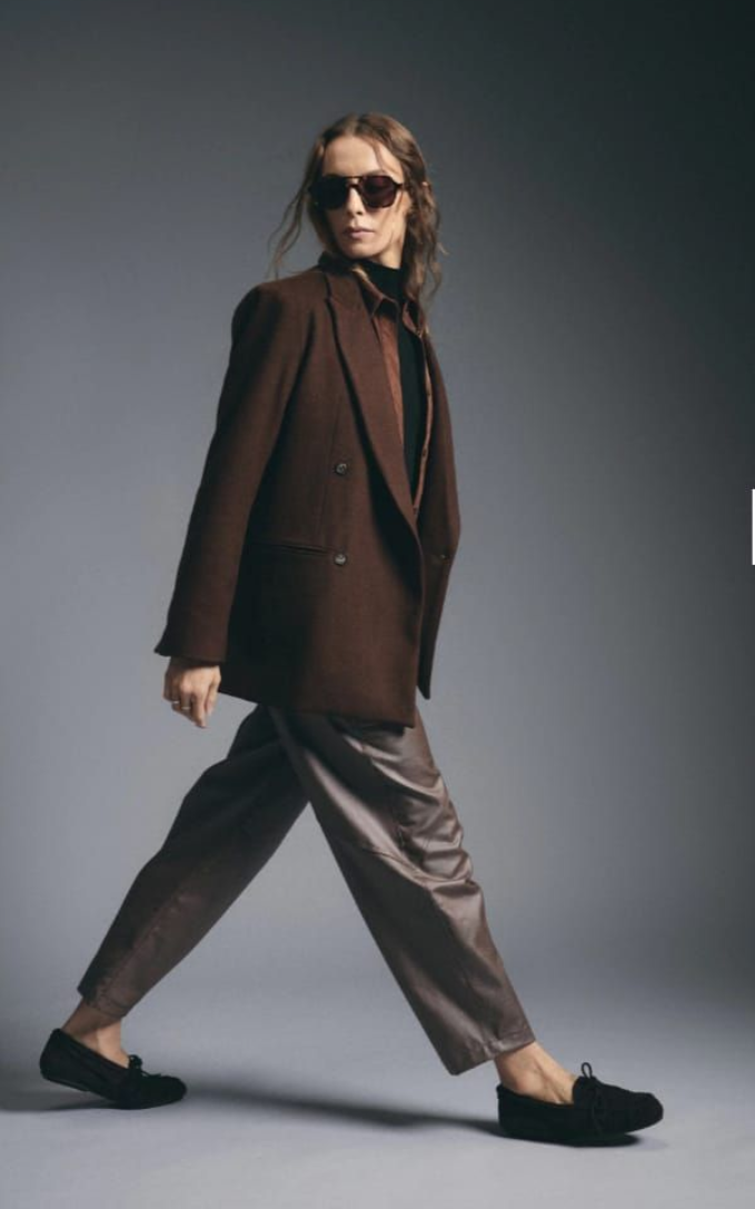 Persona con chaqueta oscura y pantalones de satén caminando de perfil contra un fondo gris de estudio.