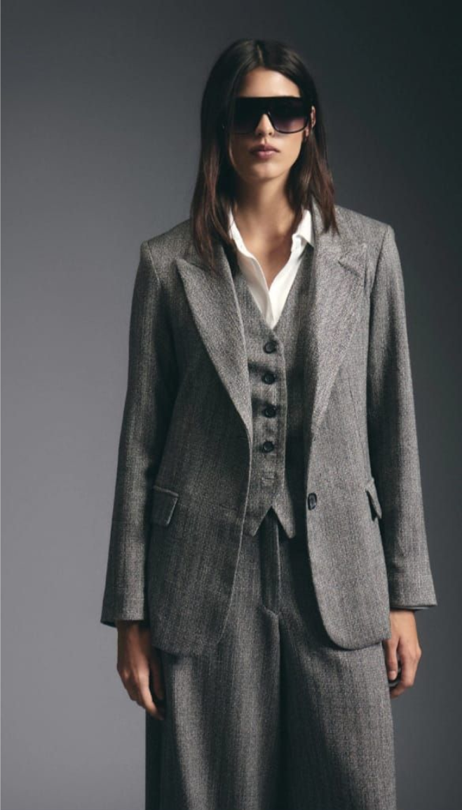 Persona con traje gris y gafas de sol ajustándose la chaqueta sobre un fondo oscuro.