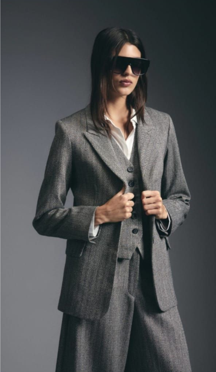 Persona con traje gris y gafas de sol ajustándose la chaqueta sobre un fondo oscuro.