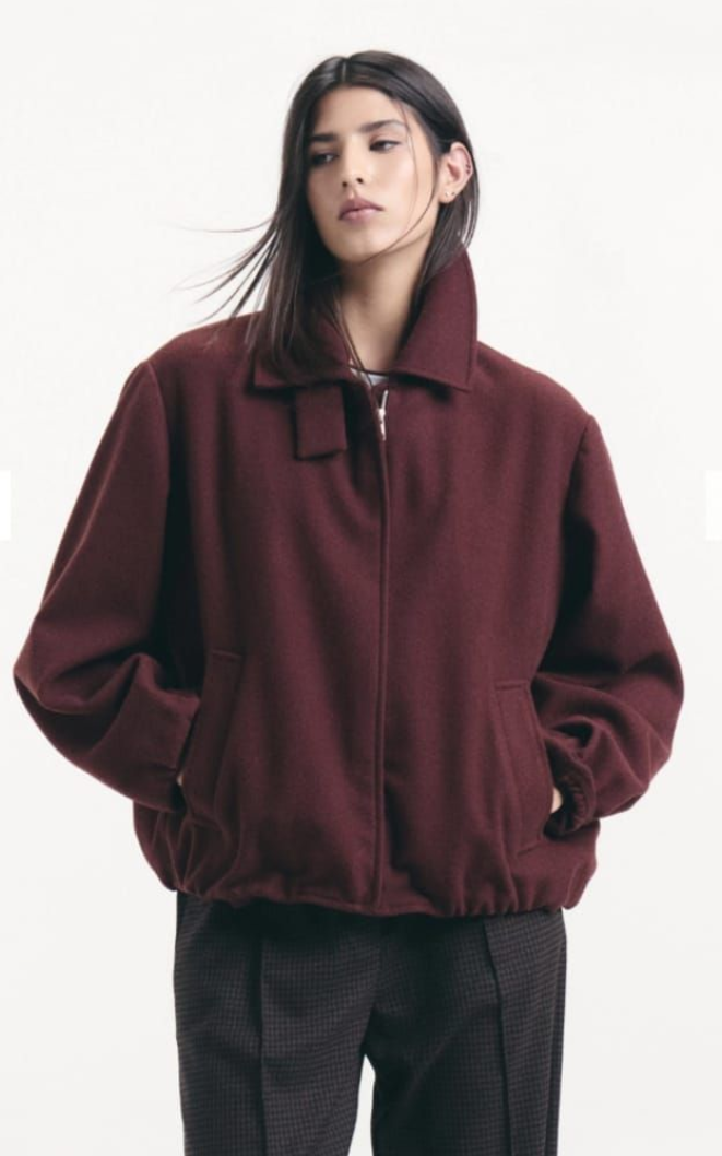 Persona que viste una chaqueta color burdeos y pantalones oscuros, de pie frente a un fondo claro liso.