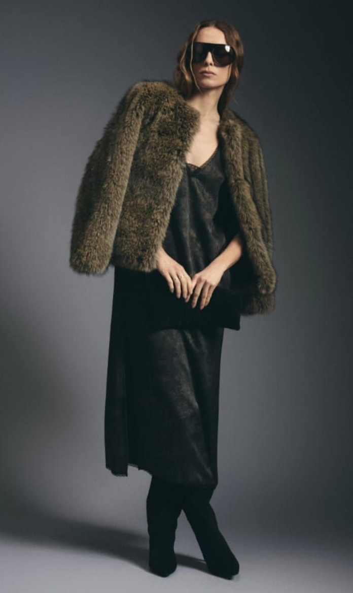 Modelo con vestido oscuro, chaqueta de piel y gafas de sol posando frente a un fondo de estudio gris.