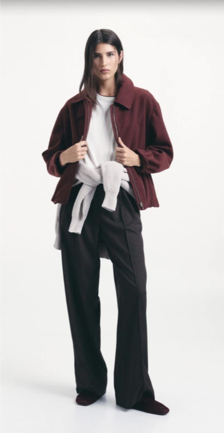 Persona que viste una chaqueta color burdeos y pantalones oscuros, de pie frente a un fondo claro liso.