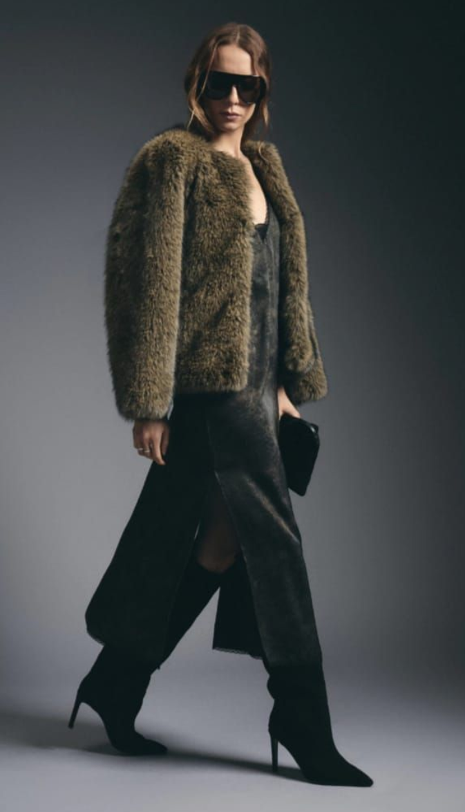 Modelo con vestido oscuro, chaqueta de piel y gafas de sol posando frente a un fondo de estudio gris.
