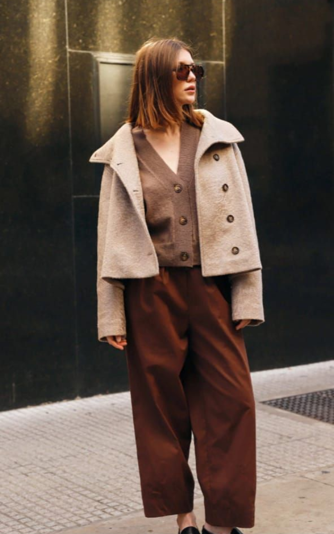 Mujer de pie en una acera de la ciudad, vestida con una chaqueta beige, pantalones marrones, gafas de sol y mocasines negros.