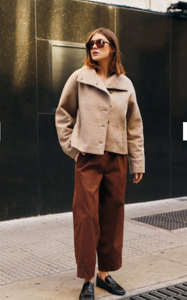 Mujer de pie en una acera de la ciudad, vestida con una chaqueta beige, pantalones marrones, gafas de sol y mocasines negros.