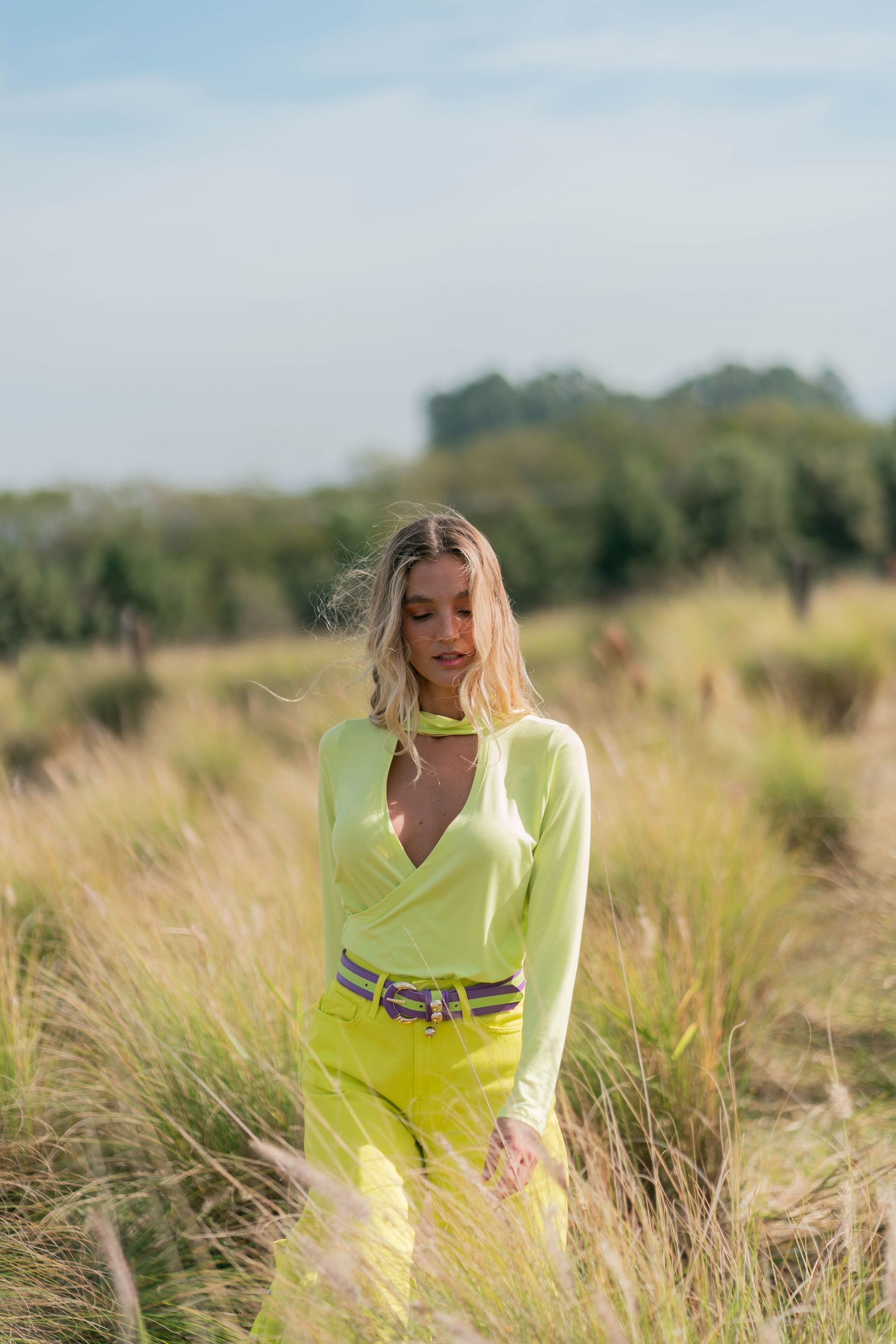 Una persona con un atuendo amarillo neón está de pie entre hierba alta con árboles al fondo.