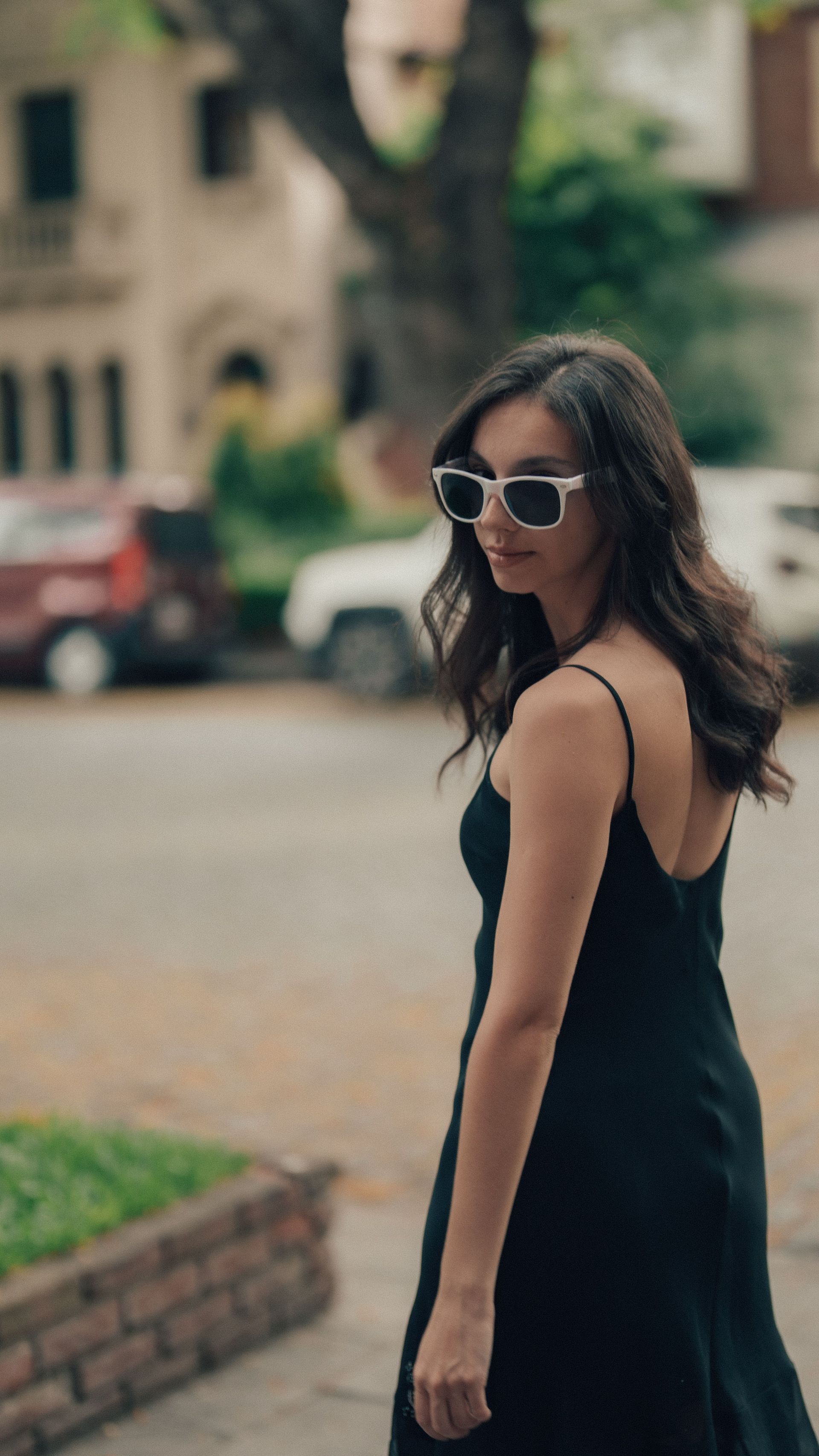 Una mujer con un vestido negro y gafas de sol está de pie en la acera, mirando por encima del hombro.
