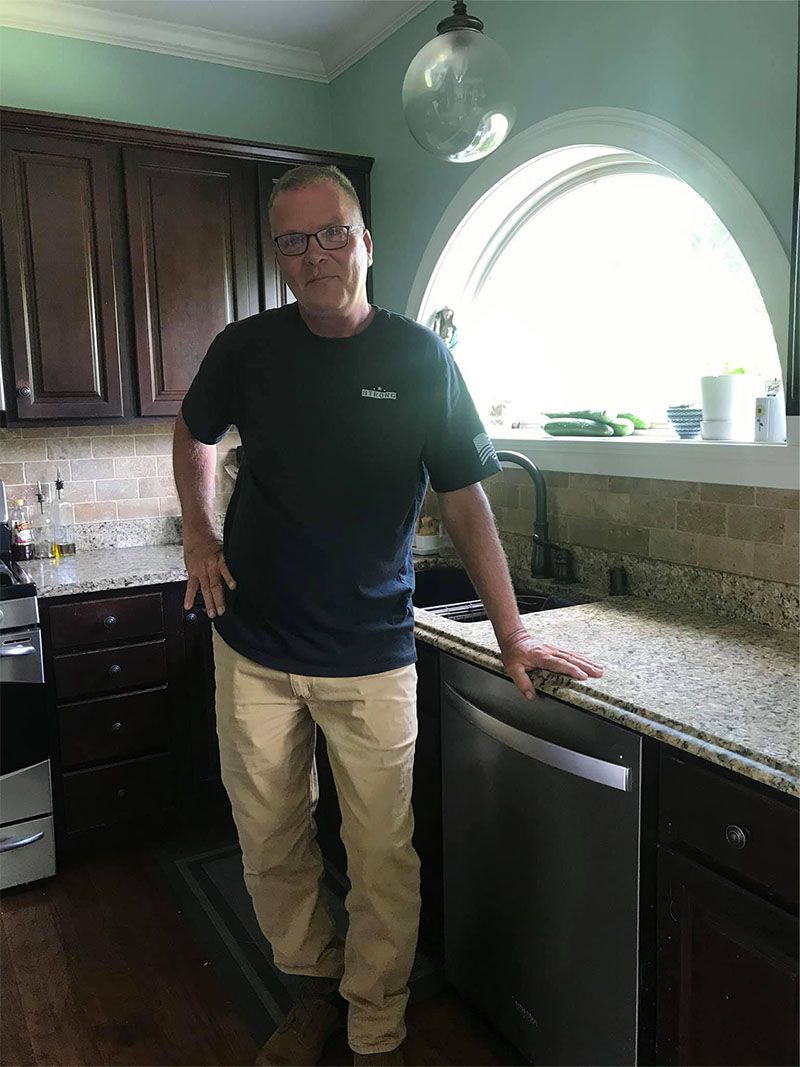 Man in a kitchen wearing a navy blue shirt and khaki pants, leaning on the countertop near the dishwasher.