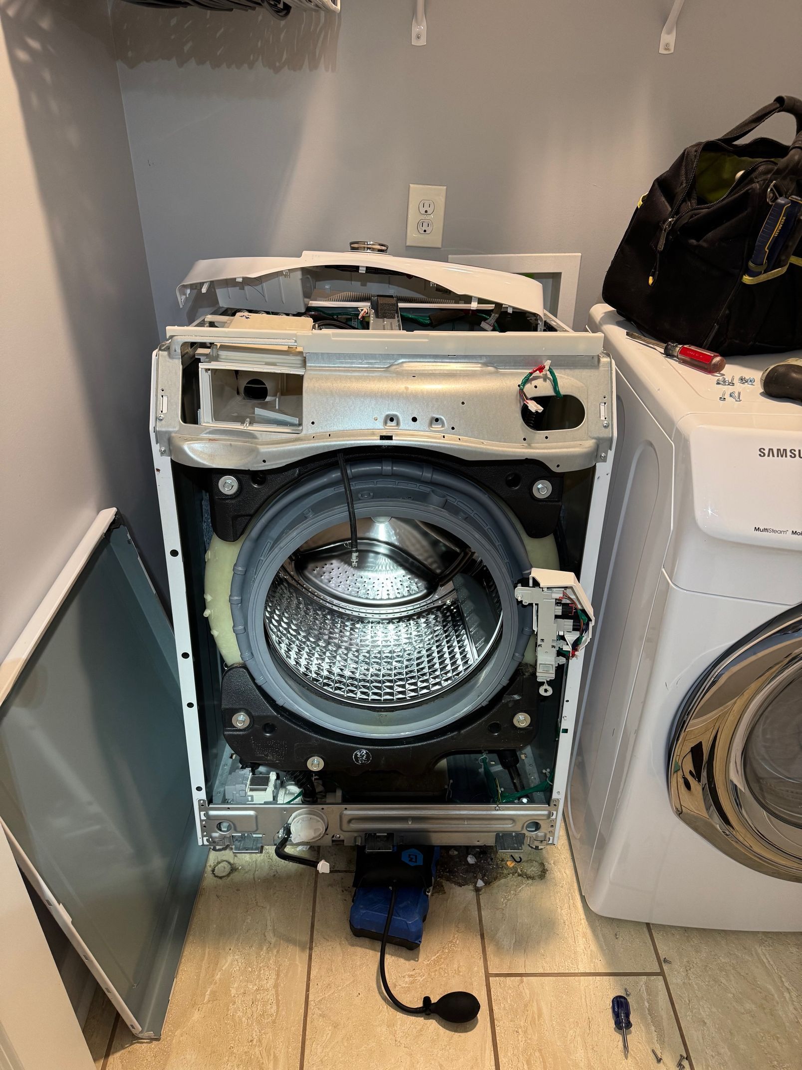 Washing machine with its front panel removed, revealing the drum and internal components. Another washer is beside it.