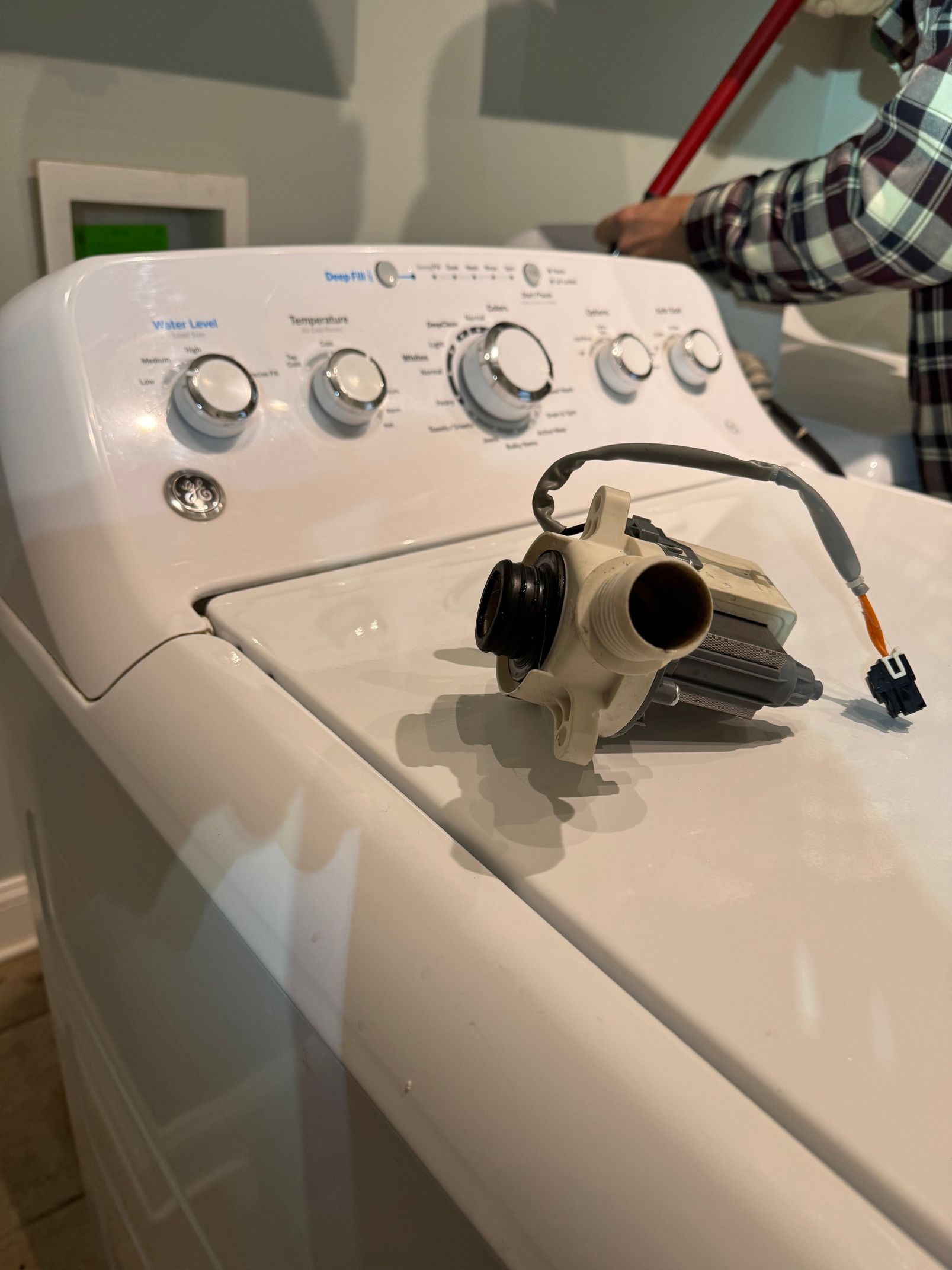 Washing machine with its pump removed, placed on top. A person's arm and a red pole are in the background.
