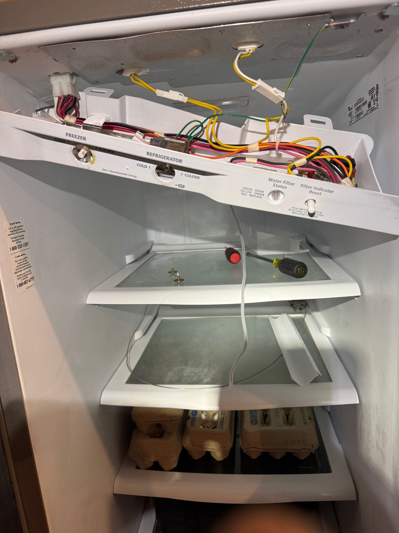 Interior of a refrigerator with shelves, lighting, and exposed wiring. A screwdriver rests on a shelf.