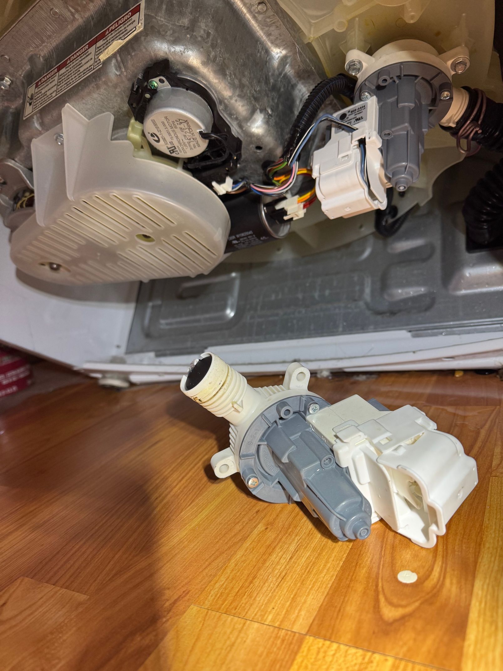 Washing machine parts: a detached pump with a grey and white motor sitting on a wooden floor near the machine's interior.