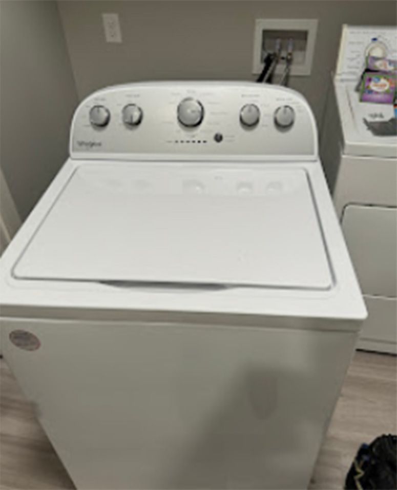 White Whirlpool washing machine in a laundry room. Controls on top, lid closed.