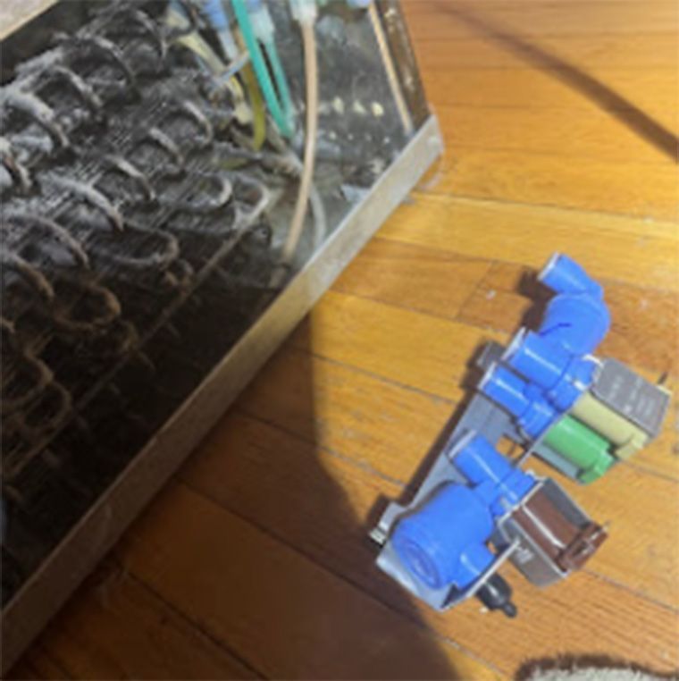 Refrigerator parts on wooden floor next to evaporator coils. Two solenoid valves are present.