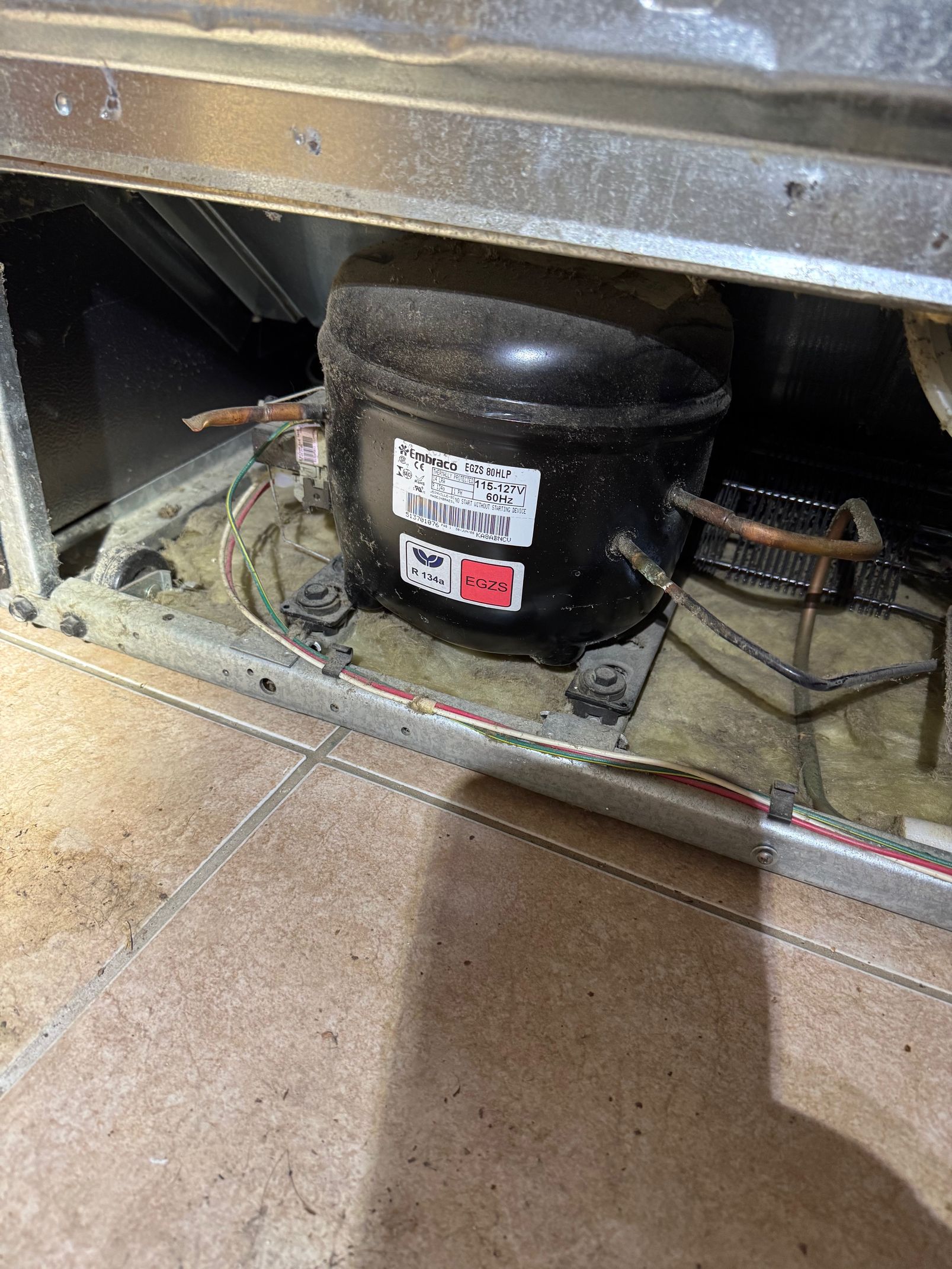 Black refrigerator compressor with copper tubing, nestled on insulation, viewed from below.