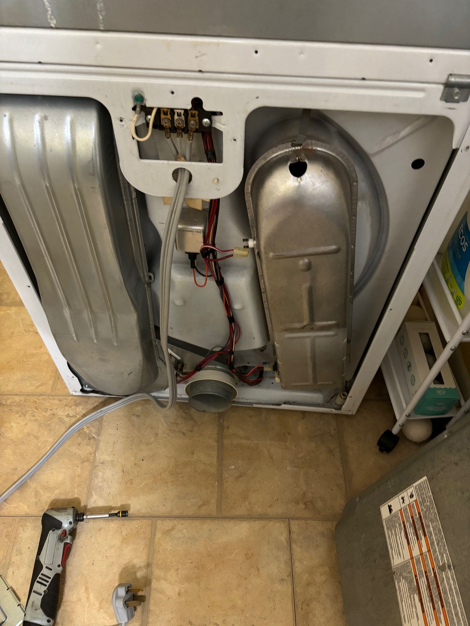 Interior of a white dryer showing the drum, heating element, and wiring; on a tile floor.