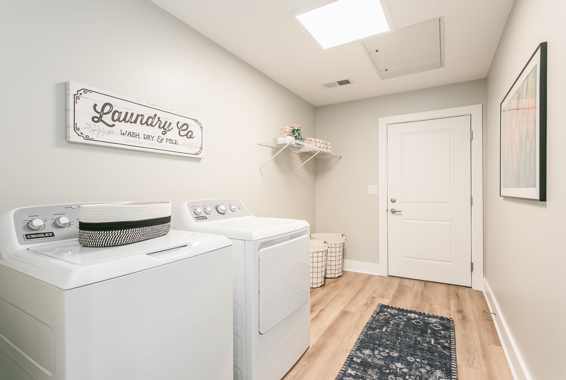 Laundry room with a washer and dryer and a closed white door. Light wooden flooring and a small rug are visible.