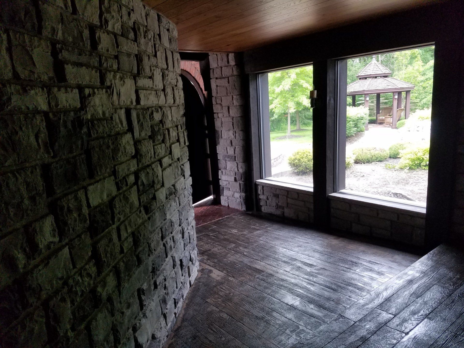 A hallway with a stone wall and wooden floor and a gazebo in the background.