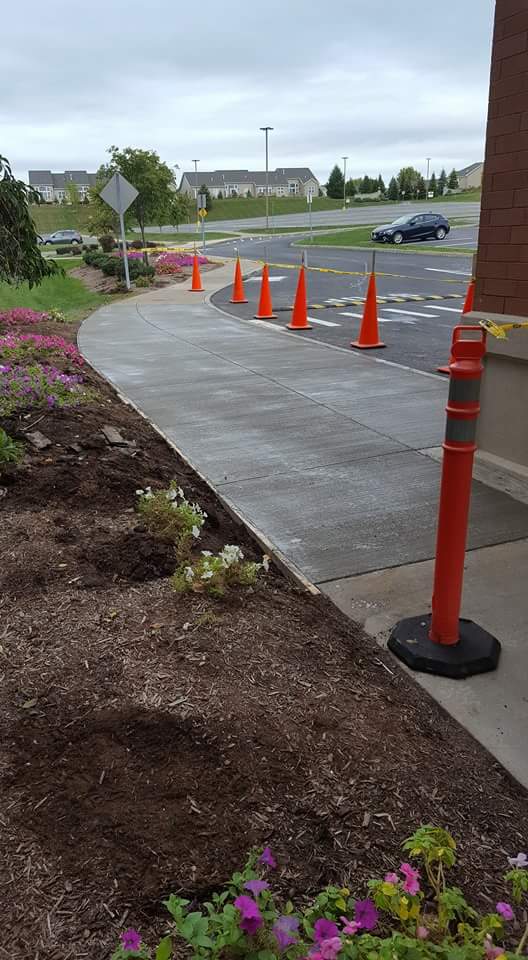 A sidewalk with orange cones on it is being built next to a parking lot.
