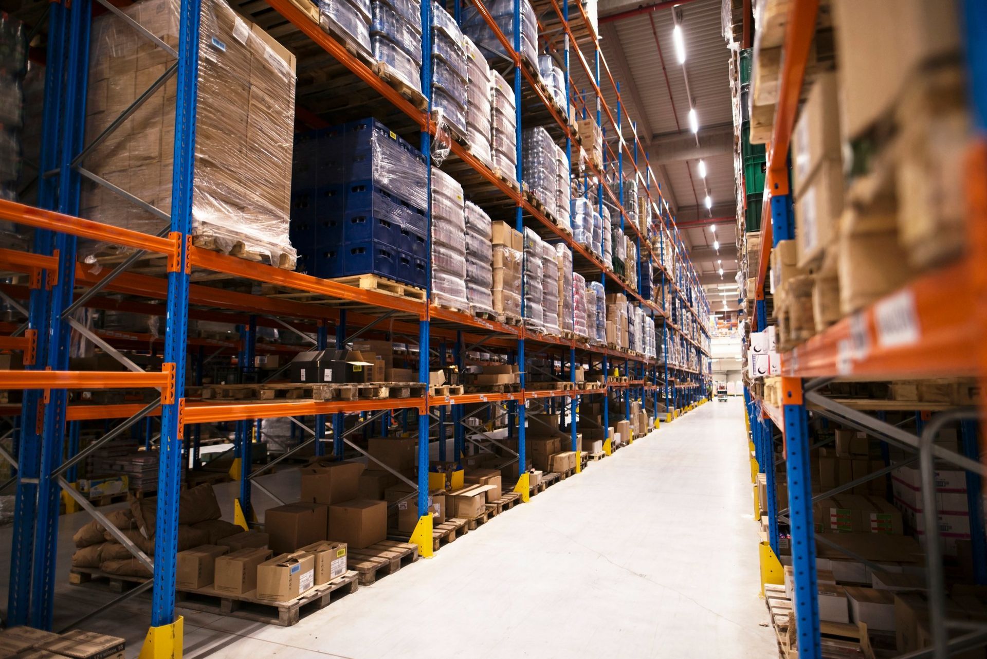 Warehouse interior with tall shelves loaded with packed goods; a wide aisle stretches between the rows.