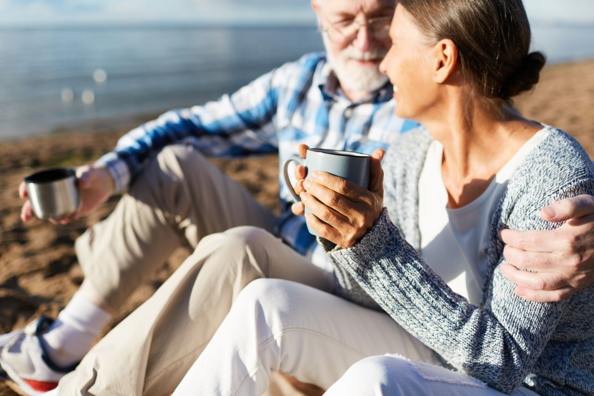 Older couple smiling and enjoying coffee on a sunny beach. Man has arm around woman.