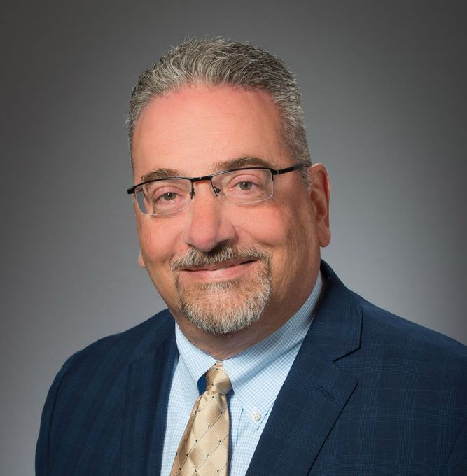 Man in a blue suit, glasses, and tie, smiling against a gray backdrop.